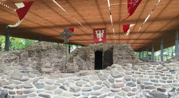 Kamienne ruiny na Ostrowiu Lednickim, z historycznymi chorągwiami i krzyżem. Wyobraź sobie, że to miejsce, skąd wyruszał dawny ostrów lednicki prom.