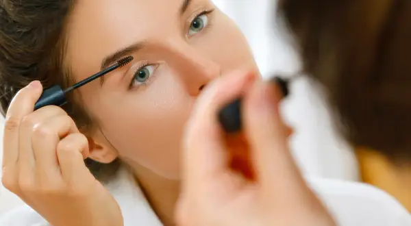 Une femme applique du mascara sur ses sourcils, préparant son maquillage bagage cabine pour un voyage.
