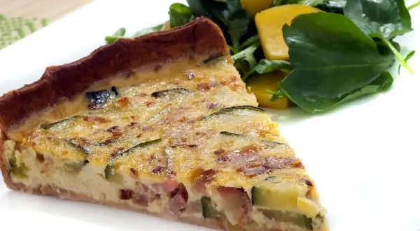 Rebanada de quiche de calabacín y bacon, con trozos de queso y una ensalada verde al lado.
