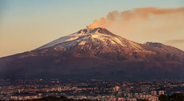 Etna, najwyższy wulkan Europy, dymi na tle zachodzącego słońca. U jego stóp rozciąga się miasto.