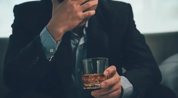 Mężczyzna w garniturze trzyma szklankę z whisky, opierając czoło na dłoni. Może to symbolizować problemy, stres, a nawet **renta alkoholowa**.