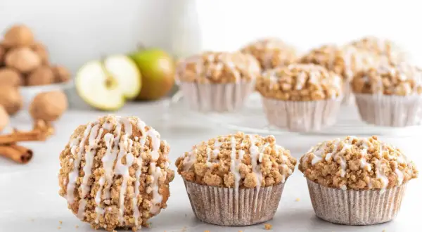 Leckere Apfelmuffins mit Streuseln und Zuckerguss, perfekt für ein Apfelmuffins Rezept. Daneben liegen Äpfel und Zimtstangen.
