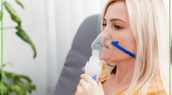 Kobieta z blond włosami używa nebulizatora, co może być związane z leczeniem mukowiscydozy.