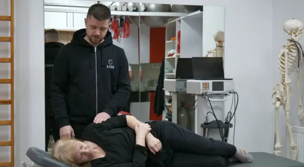 Fizjoterapeuta pomaga pacjentce, która cierpi na choroby od dźwigania. W tle model szkieletu i sprzęt rehabilitacyjny.
