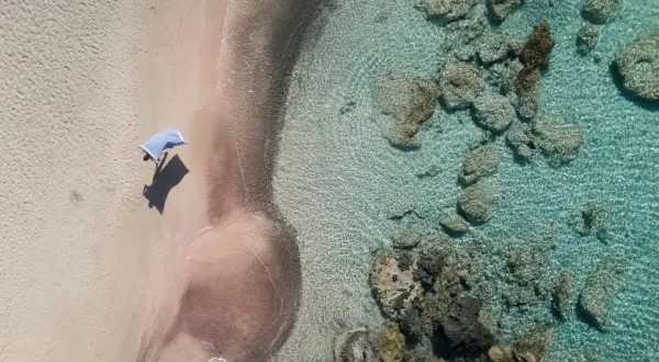En person med en blå handduk på en rosa strand på Kreta. Klart turkost vatten med stenar.
