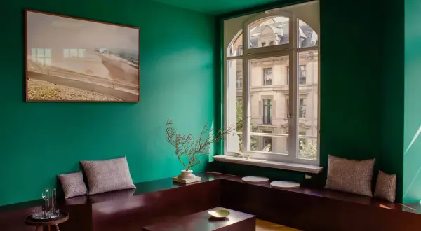 Un salotto con pareti verde scuro, un quadro paesaggistico, sedute in legno scuro e cuscini a quadri. I colori da abbinare al verde scuro includono il marrone del legno e le sfumature neutre dei cuscini.