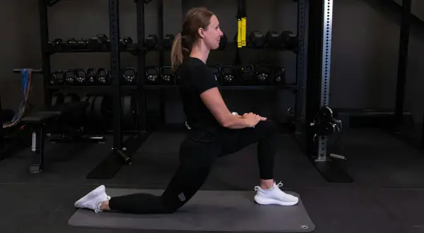 Kvinna utför grodan stretch på en träningsmatta i ett gym. Hantlar och vikter syns i bakgrunden.