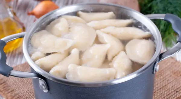 Gotowanie pierogów w garnku. Woda wrze, a pierogi unoszą się na powierzchni. W tle widać olej i koperek. Dowiedz się, ile gotować pierogi, by były idealne.