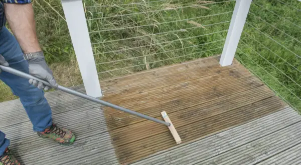 Mann reinigt mit einem Besen die Holzterrasse. Die eine Hälfte ist schon nass und dunkler, die andere noch trocken.