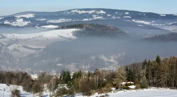 Mgła unosi się nad zimową doliną, ukazując sporą ilość pyłów zawieszonych w powietrzu. W oddali widać ośnieżone wzgórza i lasy.