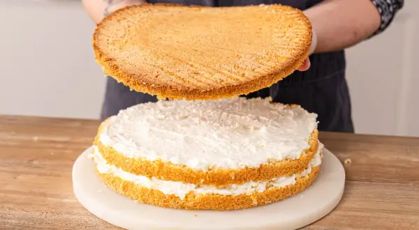 Eine Biskuit-Schicht wird auf eine mit weißer Creme gefüllte Erdbeer-Raffaello-Torte gesetzt.