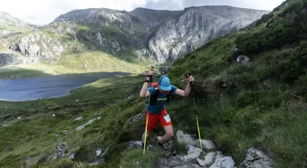 Un coureur grimpe une pente rocheuse, bâtons en main, lors de la kat100 by utmb. Un lac et des montagnes majestueuses en arrière-plan.