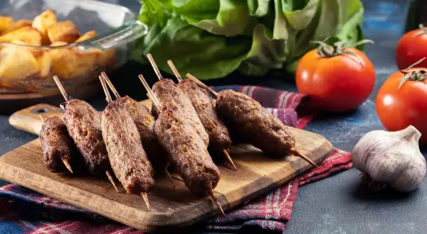 Aksamitne cevapcici z grilla, podane na desce z dodatkami: pieczone ziemniaki, pomidory i czosnek.