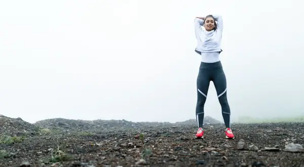 Femme en tenue de sport s'étirant sur un terrain rocailleux, prête pour son entrainement course à pied dans un paysage brumeux.