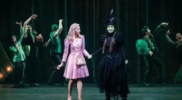 Elphaba w czarnej sukni i Glinda w różowej sukience trzymają się za ręce na scenie.