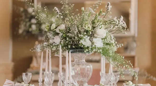 Eleganckie ustawienie stołów na 20 osób. Centralny punkt stanowi bukiet białych kwiatów w szklanym wazonie, otoczony świecami i kryształowymi kieliszkami.
