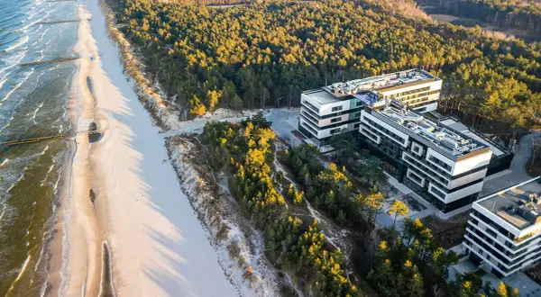 Nowoczesne budynki hotelowe w Rogowie nad morzem, otoczone sosnowym lasem, z widokiem na piaszczystą plażę i fale Bałtyku.
