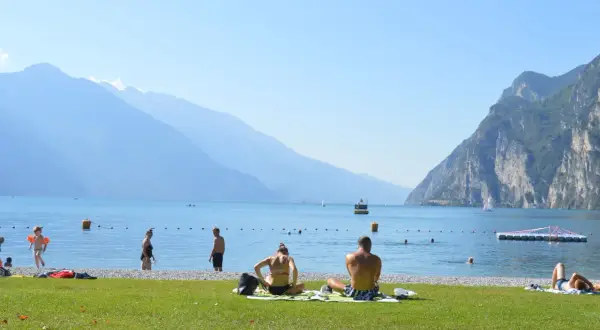Gente in spiaggia e in acqua, con montagne sullo sfondo, in una giornata di sole sul lago di Garda, forse vicino a Lazise o Bardolino.