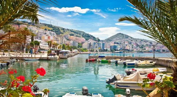 Albania, gdzie najpiękniej! Widok na port z łodziami i kolorowe miasto na wzgórzu, otoczone palmami.