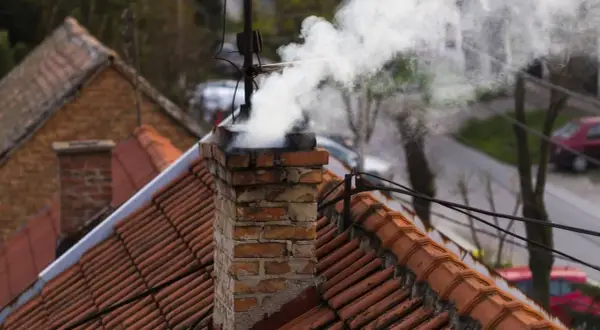 Rauch steigt aus einem alten Ziegelschornstein auf. Die Ziegel sind abgenutzt, was auf eine mögliche Schornsteinversottung durch Regen hindeutet.