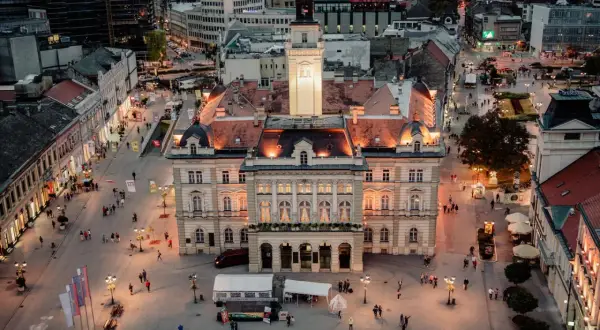 Abendliche Ansicht des Hauptplatzes in Novi Sad, Serbien, mit beleuchteten Gebäuden und Menschen, die spazieren gehen.