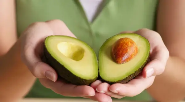 Manos sostienen un aguacate cortado por la mitad, mostrando su pulpa verde y el hueso. Así es qué es el aguacate, una fruta cremosa y nutritiva.