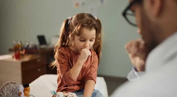 Niña con tos perruna en consulta médica. Un doctor la examina mientras ella se cubre la boca.