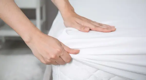 Hands smoothing a white fitted sheet over a mattress. Is it ok to change bed sheets once a month? Probably not for optimal hygiene.