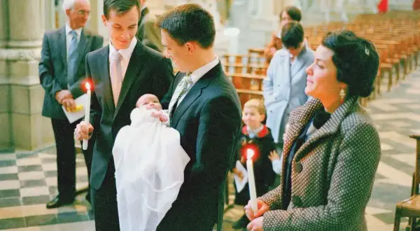 Le parrain tient sa nièce, un bébé en habit blanc, lors d'une cérémonie religieuse. Une bougie allumée est tenue par un autre homme.
