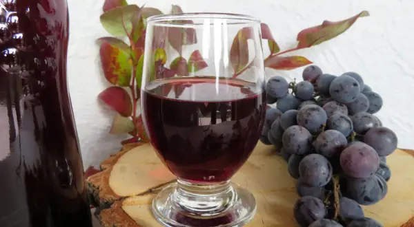 Ciemnoczerwone wino w kieliszku, obok kiść winogron i butla. Idealne do domowego wina w 5 litrowej butli.