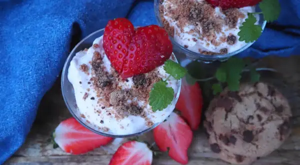 Två glas med jordgubbscheesecake, toppade med färska jordgubbar och smulor. En chokladkaka och jordgubbsbitar ligger bredvid.