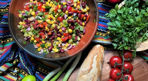 Bunter mexikanischer Salat mit Mais, Bohnen und Gemüse in einer Schüssel, daneben Baguette, Limette und Kräuter.