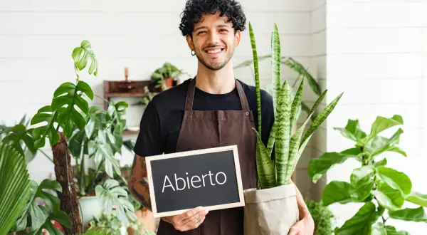Joven sonriente con plantas, sosteniendo un cartel de "Abierto". Inspiración para ideas de negocios para jóvenes en el sector verde.