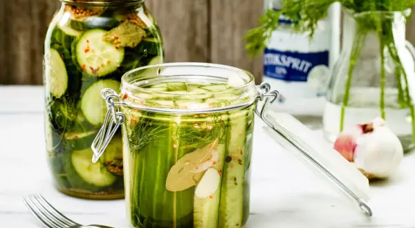 Två glasburkar fyllda med krispig, picklad gurka, dill och kryddor. En gaffel ligger bredvid.