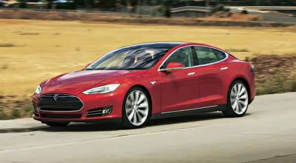 Czerwony Tesla Model S pędzi po drodze, otoczony suchymi, trawiastymi krajobrazami.