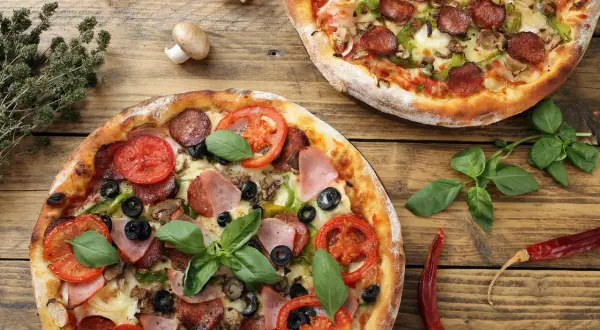Dwie pizze z salami, pomidorami, oliwkami, szynką i bazylią. Widać jakie składniki na pizze można wybrać.