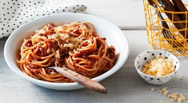 En skål med stuvad spaghetti med köttfärssås och riven ost. En liten skål med mer ost och en korg med bestick står bredvid.
