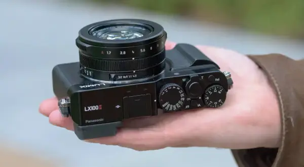 A hand holds a Panasonic Lumix DMC-LX100K camera, showcasing its compact design and prominent lens.