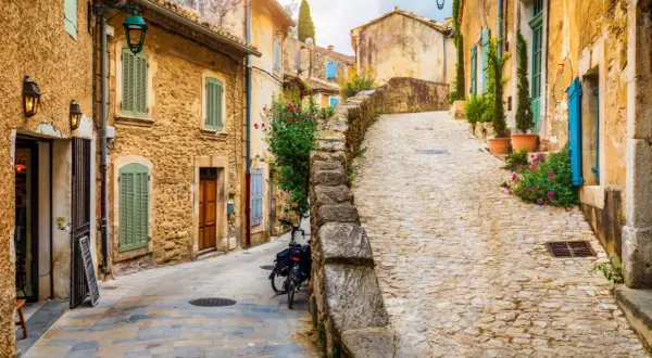 Ruelle pavée d'un village du Luberon, maisons en pierre ocre, volets verts et bleus, fleurs colorées. Parfait pour un week-end Luberon.