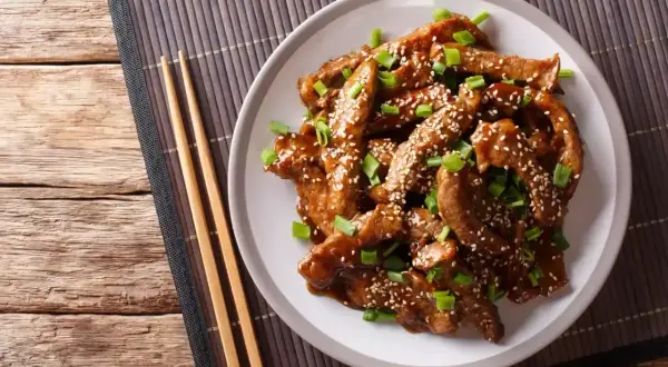 Pyszny kurczak teriyaki z sezamem i szczypiorkiem na talerzu, obok pałeczki.