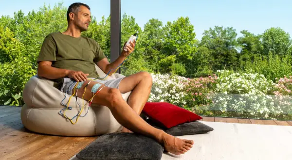 Homme assis, appliquant des électrodes sur sa cuisse, appareil de stimulation en main. Il observe le paysage fleuri, sans effets secondaires apparents du urostim.