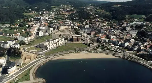 Vista aérea de la playa de Cee, con el mar azul, la arena dorada y la ciudad extendiéndose hacia las colinas verdes.