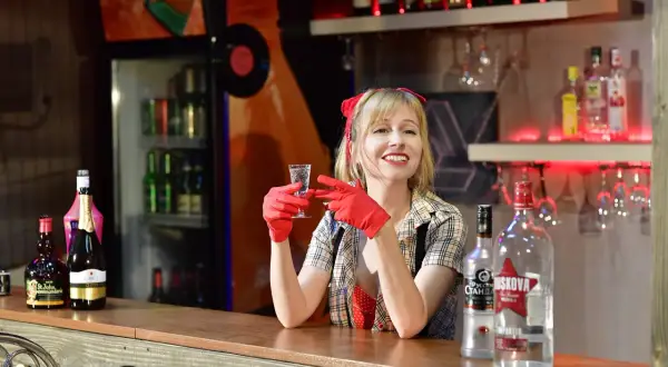 Uśmiechnięta barmanka w czerwonych rękawiczkach trzyma kieliszek z wódką. Obok stoją butelki, w tym duża 250 ml wódki.