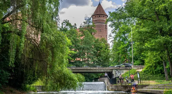 Malownicza przystań w Olsztynie z wieżą zamkową w tle. Woda, zieleń i ludzie odpoczywający nad rzeką tworzą spokojny krajobraz.