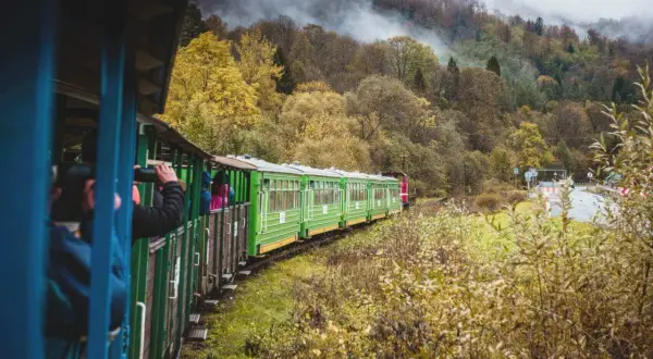 Zielony pociąg turystyczny wjeżdża w jesienne Bieszczady. Mgła spowija wzgórza, a liście mienią się złotem.
