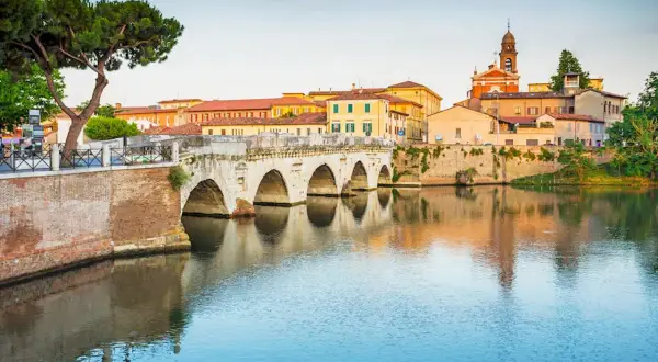 Il Ponte di Tiberio a Rimini, con i suoi archi in pietra che si specchiano sull'acqua calma, fa da sfondo a un paesaggio urbano colorato.