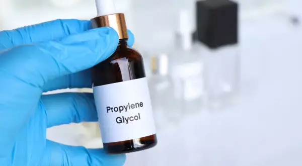 Ręka w niebieskiej rękawiczce trzyma buteleczkę z etykietą "Propylene Glycol".