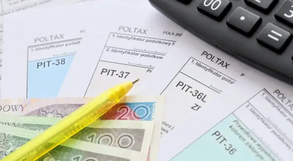 Formularze podatkowe PIT, polskie banknoty, żółty długopis i kalkulator.