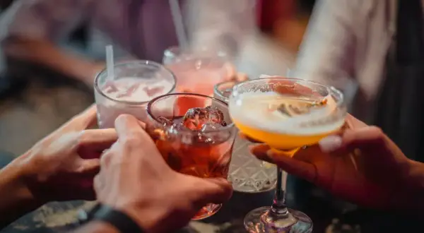 Ręce wznoszą kieliszki z kolorowymi drinkami. Wśród nich widać czerwony koktajl z lodem, różowy napój ze słomką i żółty drink w kieliszku. To idealny moment na toast z dobrym **medoc alkohol**!