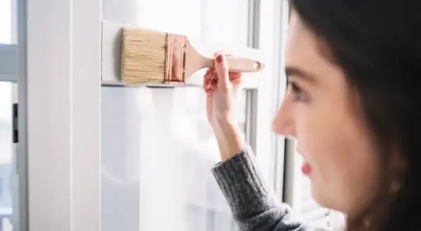 Mujer aplica pintura para persianas PVC en un marco blanco.